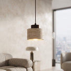 A modern cylindrical pendant light with a textured beige stone or concrete shade and a dark metal top, hanging in a softly lit living room with a window and blurred furniture in the background.