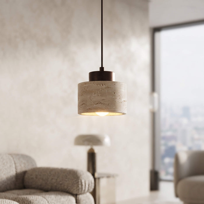 A modern cylindrical pendant light with a textured beige stone or concrete shade and a dark metal top, hanging in a softly lit living room with a window and blurred furniture in the background.