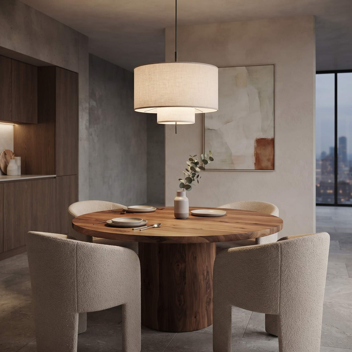 A modern dining room scene featuring a round wooden dining table with four upholstered dining chairs, a double-tiered drum pendant light, and a neutral-toned abstract wall art piece.