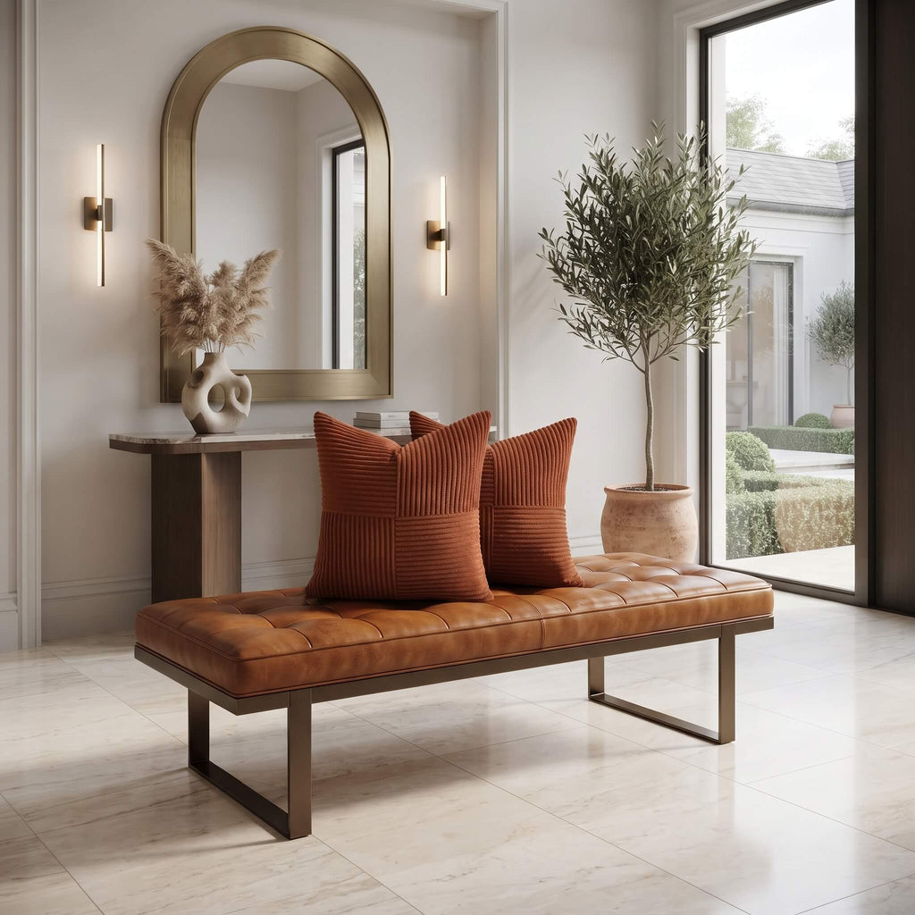 A modern tan leather tufted bench with dark metal legs in a bright, contemporary living room, styled with two rust-orange ribbed throw pillows. The room features a marble console table, a large arched gold-framed mirror, wall sconces, and a potted olive tree near large floor-to-ceiling windows.