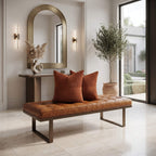 A modern tan leather tufted bench with dark metal legs in a bright, contemporary living room, styled with two rust-orange ribbed throw pillows. The room features a marble console table, a large arched gold-framed mirror, wall sconces, and a potted olive tree near large floor-to-ceiling windows.