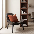 A modern curved lounge chair in dark gray with a rust-orange textured throw pillow, set in a minimalist office or study room next to a tall wooden bookshelf.