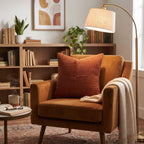 A cozy living room scene featuring a warm brown velvet armchair with a rust-orange ribbed accent pillow, a cream throw blanket, and a tall brass floor lamp in the background.