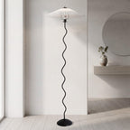 A modern floor lamp with a black wavy metal stand and a white pleated umbrella-style shade, shown in a minimalist interior room with light wood flooring and a wall shelf.