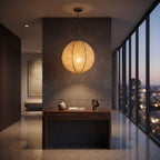 A spherical woven pendant light glowing with warm light, hanging over a dark wood console table in a modern interior hallway with large windows overlooking a city skyline at dusk.