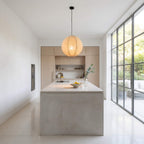 A modern kitchen island with a light gray concrete-look base and a white countertop, featuring a large woven pendant light hanging above. The background shows light wood cabinetry, a built-in sink, and a large grid-style window looking out to greenery.
