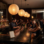 A dimly lit bar interior with warm hanging paper lanterns, a dark marble counter, and several people seated and standing around the bar. A bartender is visible behind the counter, and a menu card sits on the counter in the foreground.