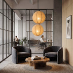 A modern interior lounge area with two dark gray armchairs facing a round wooden coffee table, set on a textured rug. Two glowing woven pendant lights hang above, and a glass partition reveals an office workspace in the background with two seated people.