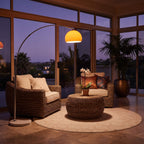 A styled outdoor patio conversation set featuring a brown wicker armchair with cream cushions, a matching loveseat with patterned pillows, a round wicker coffee table, and a large arched floor lamp with a glowing yellow globe shade, all arranged on a round beige rug in front of large glass windows at dusk.