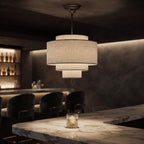 A modern three-tier pendant light with a textured beige linen shade hangs over a marble bar counter in a dimly lit bar setting.