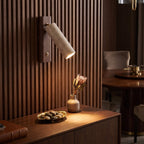 A modern wall-mounted cylindrical lamp with a light beige stone or concrete shade and a dark brown metal base, shown lit on a slatted wood wall above a wooden console table in a warm, upscale interior.