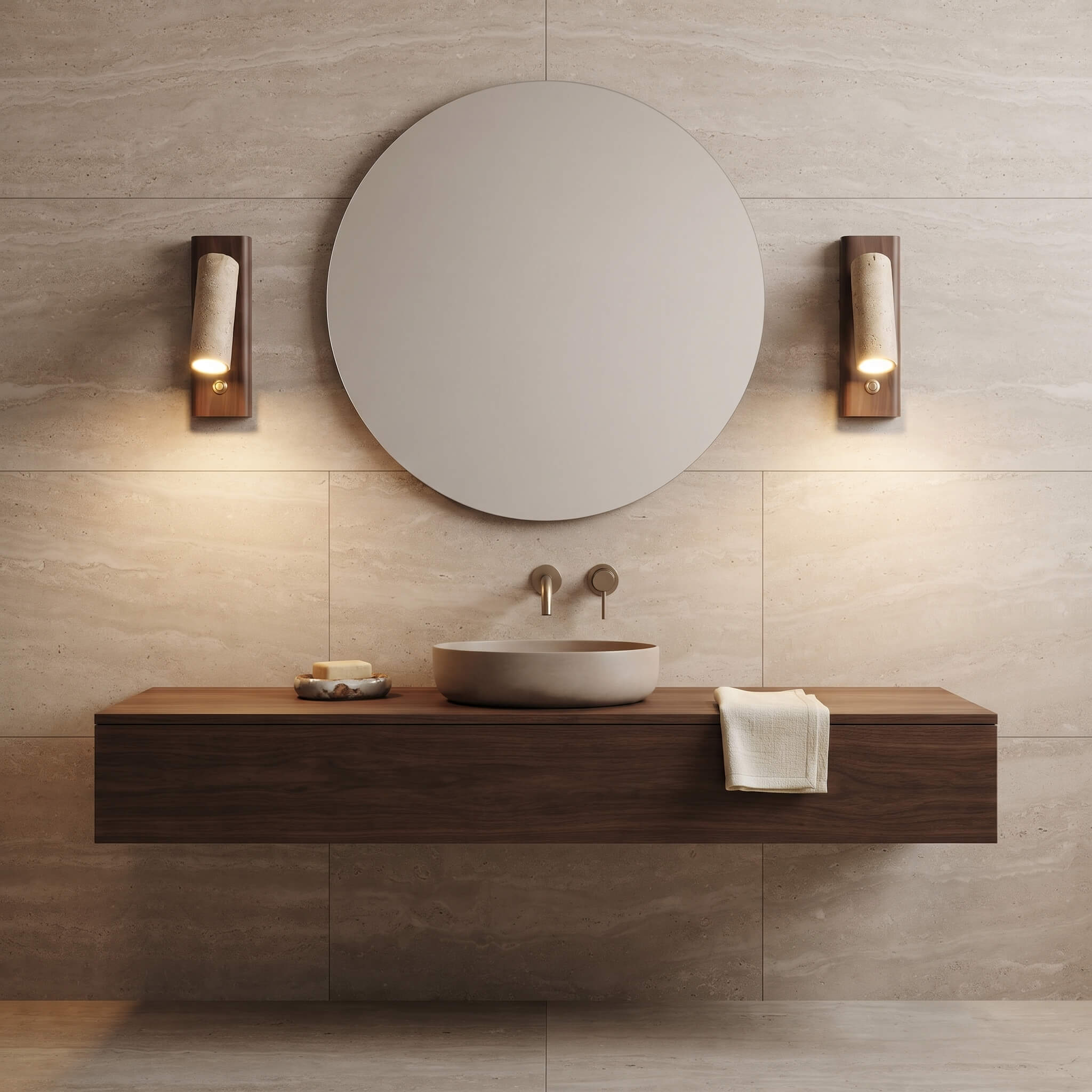 A modern floating bathroom vanity with a round vessel sink, matte wood countertop, wall-mounted brass faucets, a large round mirror, and two matching wall sconces against a light stone tile wall.