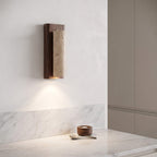 A modern wall sconce with a dark wood frame and a textured beige stone cylinder, mounted on a white wall above a marble countertop.