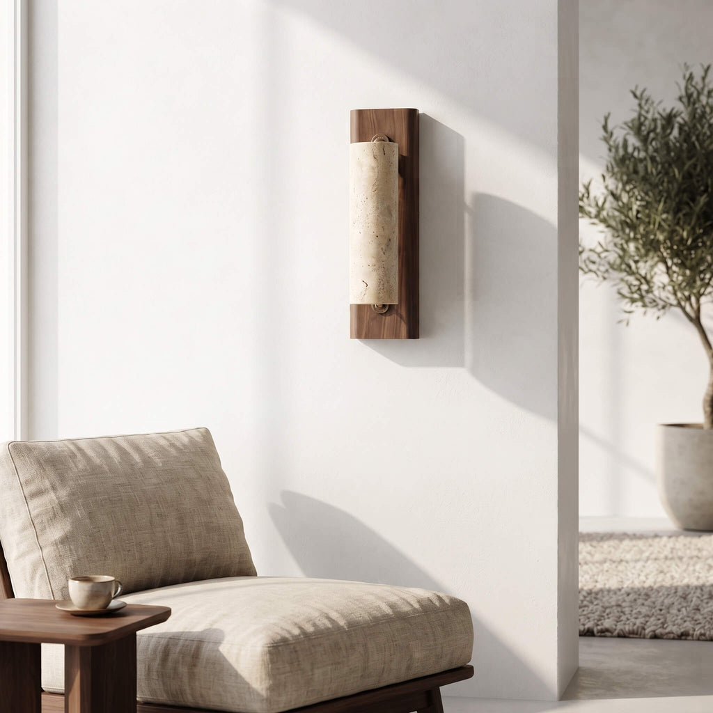 A modern wall-mounted sconce with a vertical rectangular wood frame and a textured beige fabric shade, shown mounted on a white wall in a minimalist interior setting with a beige upholstered chair and a small side table.