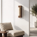 A modern wall-mounted sconce with a vertical rectangular wood frame and a textured beige fabric shade, shown mounted on a white wall in a minimalist interior setting with a beige upholstered chair and a small side table.