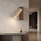 A modern wall-mounted sconce with a cylindrical textured beige shade and a dark wood backplate, casting a warm light on a beige plaster wall above a stone console table.