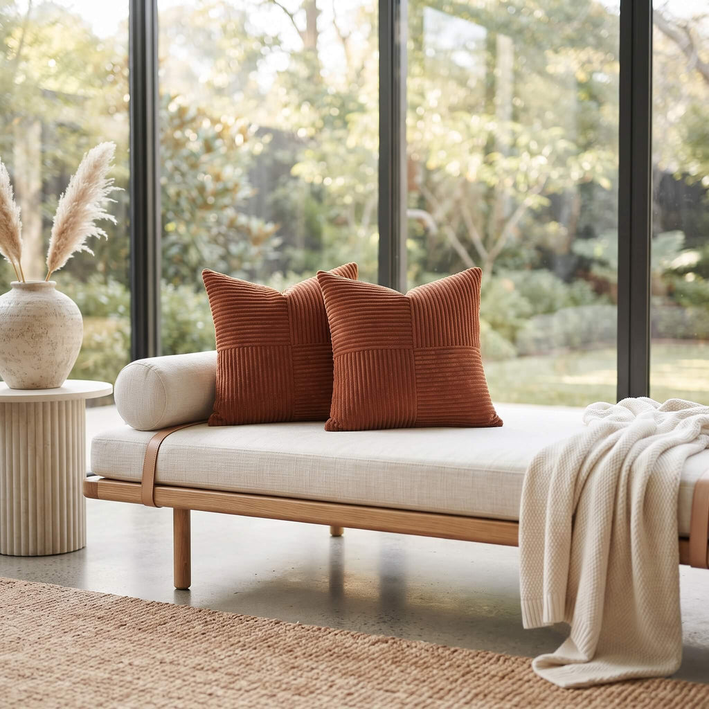 A modern outdoor daybed with a light beige linen cushion and two rust-orange ribbed throw pillows, styled with a cream knit blanket and set in a bright room with large windows overlooking greenery.