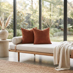 A modern outdoor daybed with a light beige linen cushion and two rust-orange ribbed throw pillows, styled with a cream knit blanket and set in a bright room with large windows overlooking greenery.