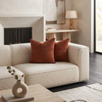 A cream boucle loveseat sofa in a modern living room setting, styled with two rust-orange ribbed throw pillows, a small ceramic vase on a wooden coffee table, and a fireplace in the background.
