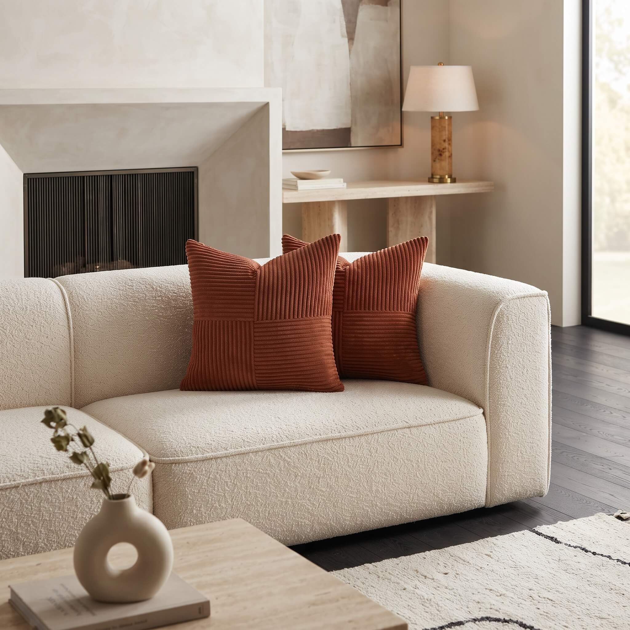 A cream boucle loveseat sofa in a modern living room setting, styled with two rust-orange ribbed throw pillows, a small ceramic vase on a wooden coffee table, and a fireplace in the background.