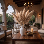 A large white ceramic vase filled with tall beige pampas grass sits on a dark wood coffee table in a luxurious, sunlit Mediterranean-style patio lounge. The setting features cream-colored armchairs, stone arches, a black chandelier, and a lit pillar candle in a glass holder.