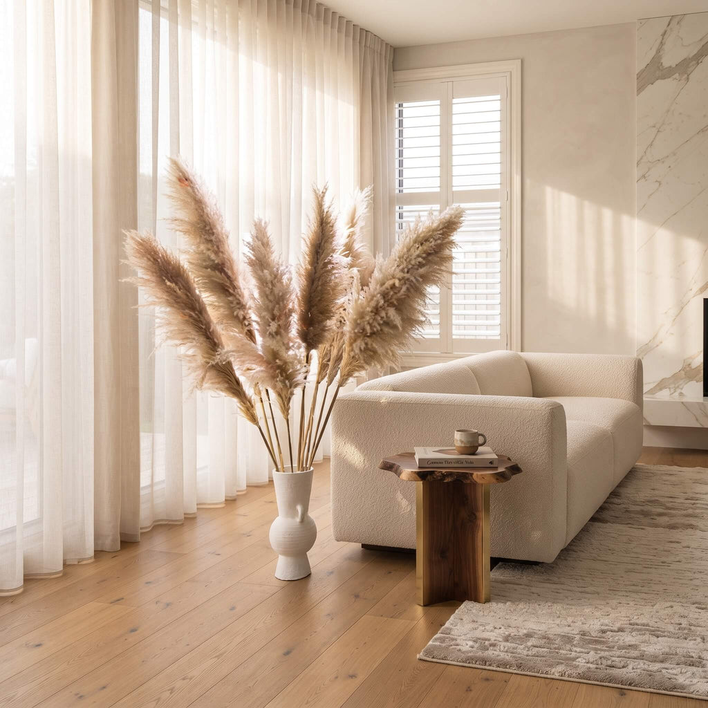 A styled living room scene featuring a cream-colored sofa, a rustic wooden side table with a small cup, a tall white vase filled with pampas grass, and a textured area rug on a light wood floor.