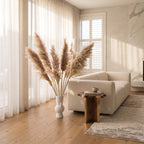 A styled living room scene featuring a cream-colored sofa, a rustic wooden side table with a small cup, a tall white vase filled with pampas grass, and a textured area rug on a light wood floor.