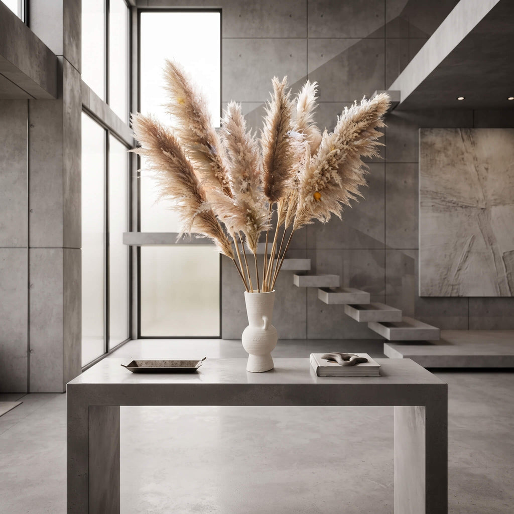 A white sculptural ceramic vase filled with tall beige pampas grass, placed on a gray concrete table in a modern minimalist interior with concrete walls, floating stairs, and large windows.
