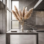 A white sculptural ceramic vase filled with tall beige pampas grass, placed on a gray concrete table in a modern minimalist interior with concrete walls, floating stairs, and large windows.
