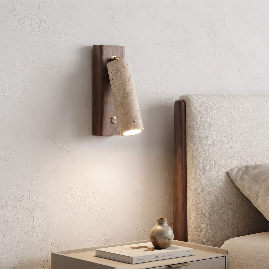 A modern wall-mounted sconce with a dark wood backplate and a tapered textured stone shade, shown lit in a bedroom setting next to a bed.