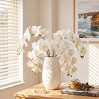 White artificial orchid flowers arranged in a matte white ceramic vase on a light wood table in a sunlit room, with a framed coastal print and small decorative objects nearby.
