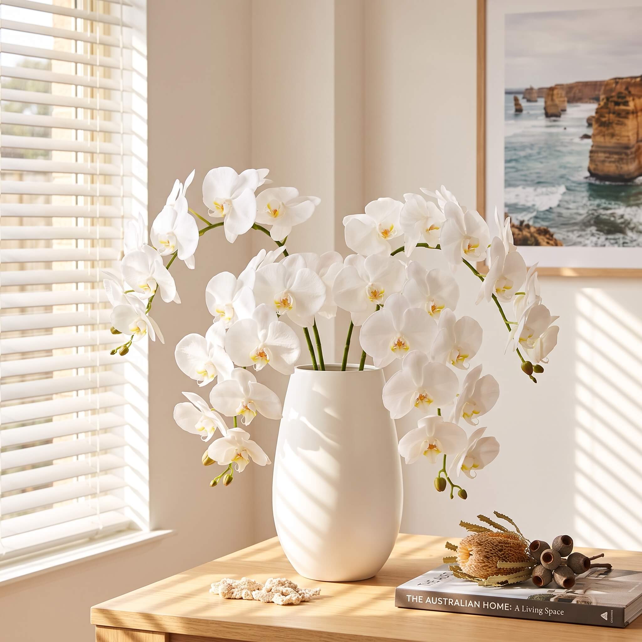 White artificial orchid flowers arranged in a matte white ceramic vase on a light wood table in a sunlit room, with a framed coastal print and small decorative objects nearby.