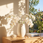 White artificial orchid flowers arranged in a matte white ceramic vase on a light wood table, with a window showing greenery outside and warm sunlight casting shadows.