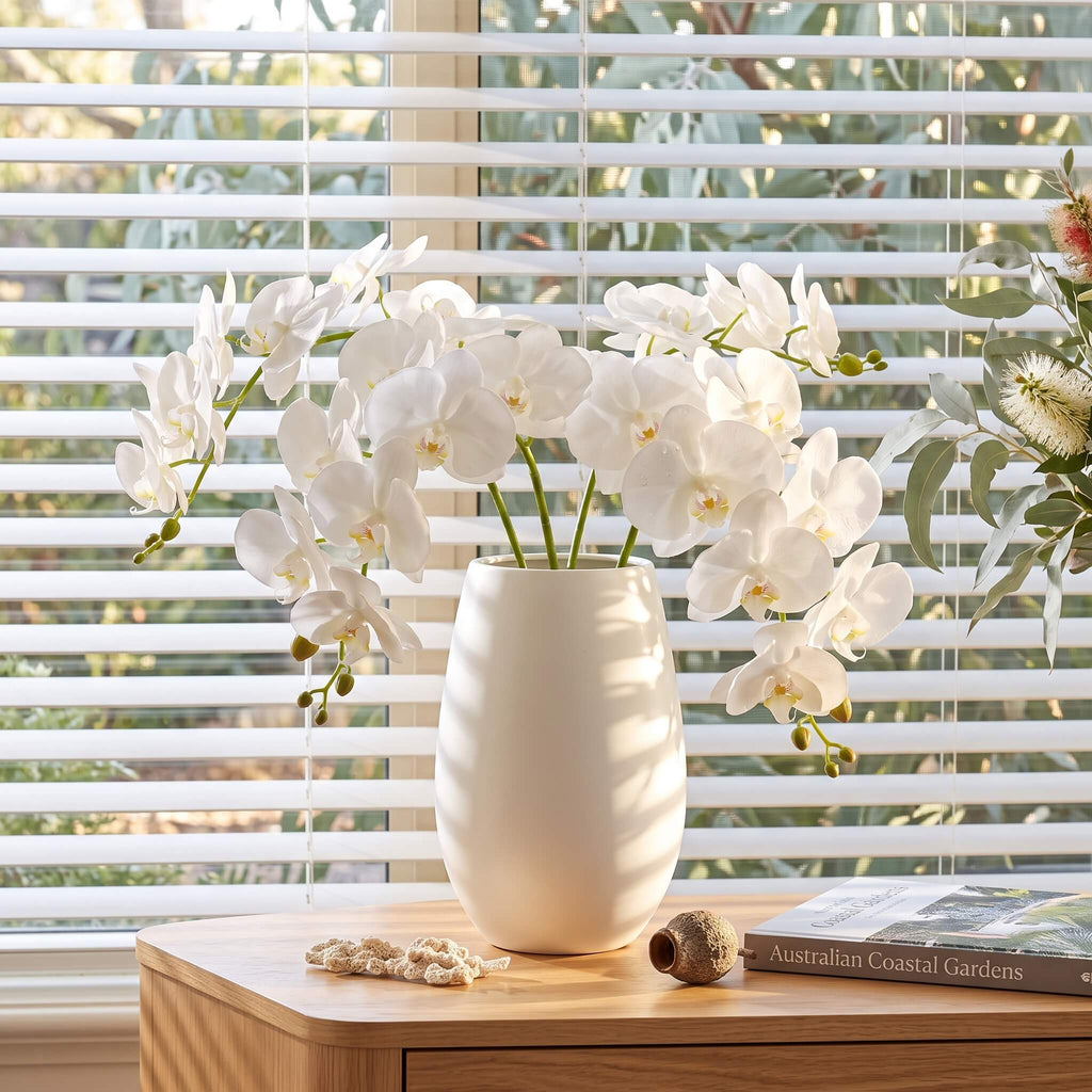 White artificial orchid flowers arranged in a matte white ceramic vase on a light wood table, set in front of white window blinds with outdoor greenery visible in the background.