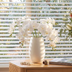 White artificial orchid flowers arranged in a matte white ceramic vase on a light wood table, set in front of white window blinds with outdoor greenery visible in the background.