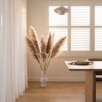 A styled interior scene with a tall vase of pampas grass beside a wooden dining table and a window with white shutters, in a warm neutral room.