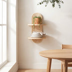 A light natural wood wall-mounted shelf with two tiers and an arched top, styled with a small potted trailing plant and stacked white plates, in a bright minimalist dining room next to a wooden table.